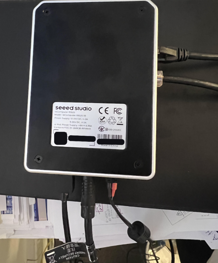 A Seeed Studio reComputer R1025-10 mini PC placed on a desk, connected with several cables. The back label displays technical specifications and certification logos, including power input details and compliance marks.
